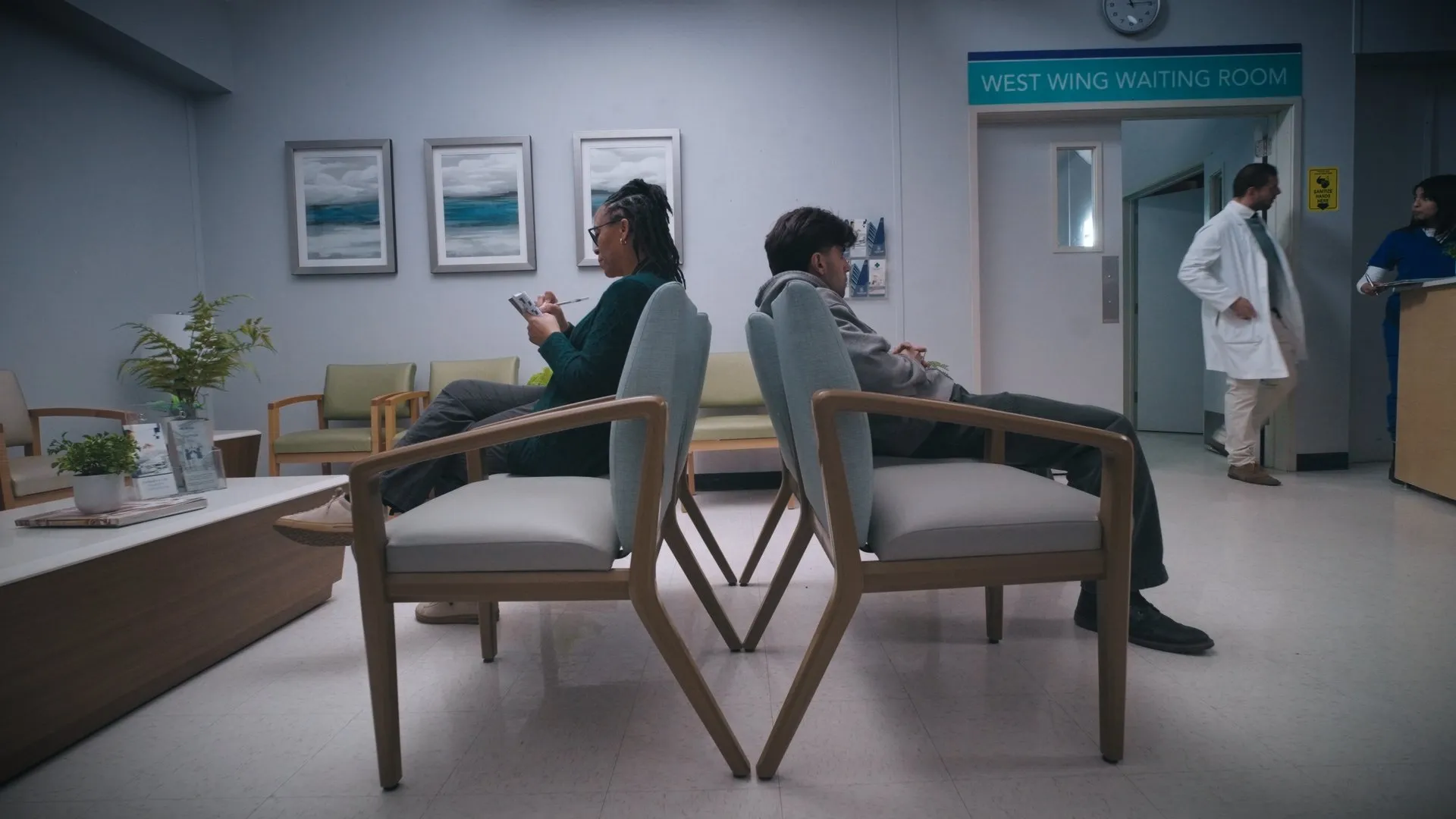 Wide shot of a quiet hospital waiting room with two people seated apart.