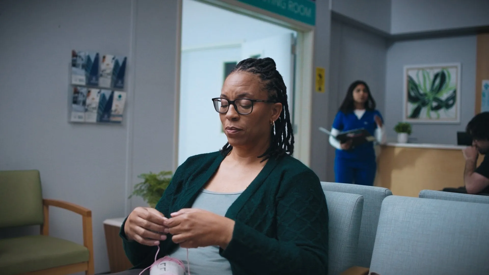 Woman in glasses knitting alone in a hospital waiting room.