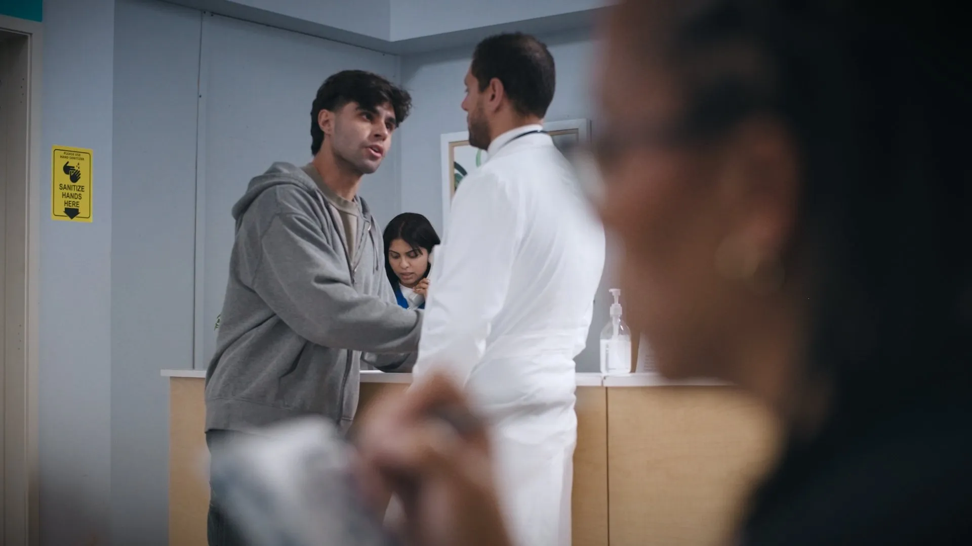 Young man in a grey hoodie speaking with a doctor at the nurses’ station.