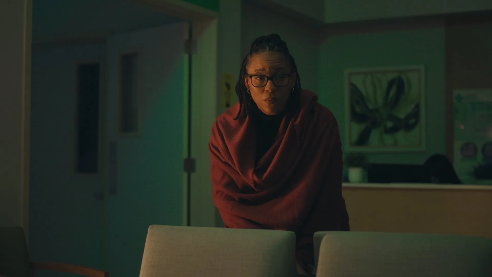 Woman wrapped in a red shawl sitting alone in a dimly lit waiting room at night.