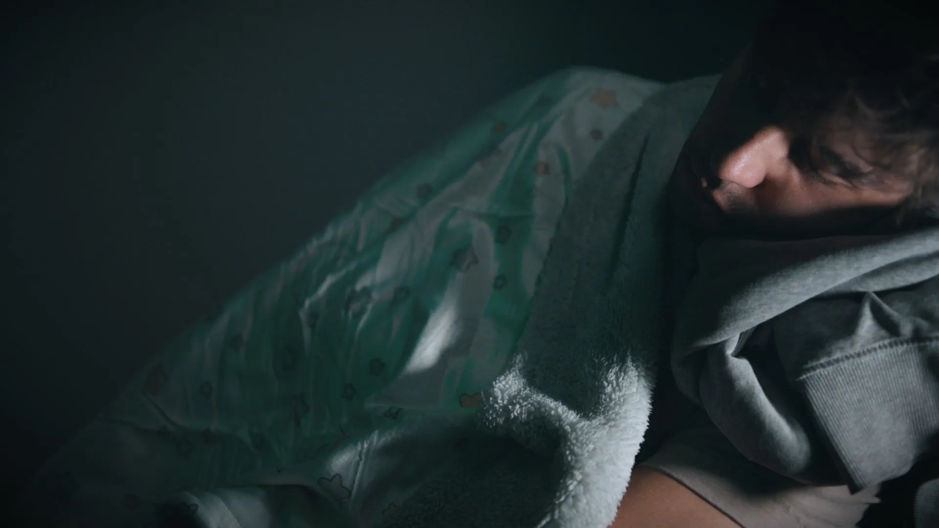 Close-up of a young man asleep, curled up under a dark green blanket.