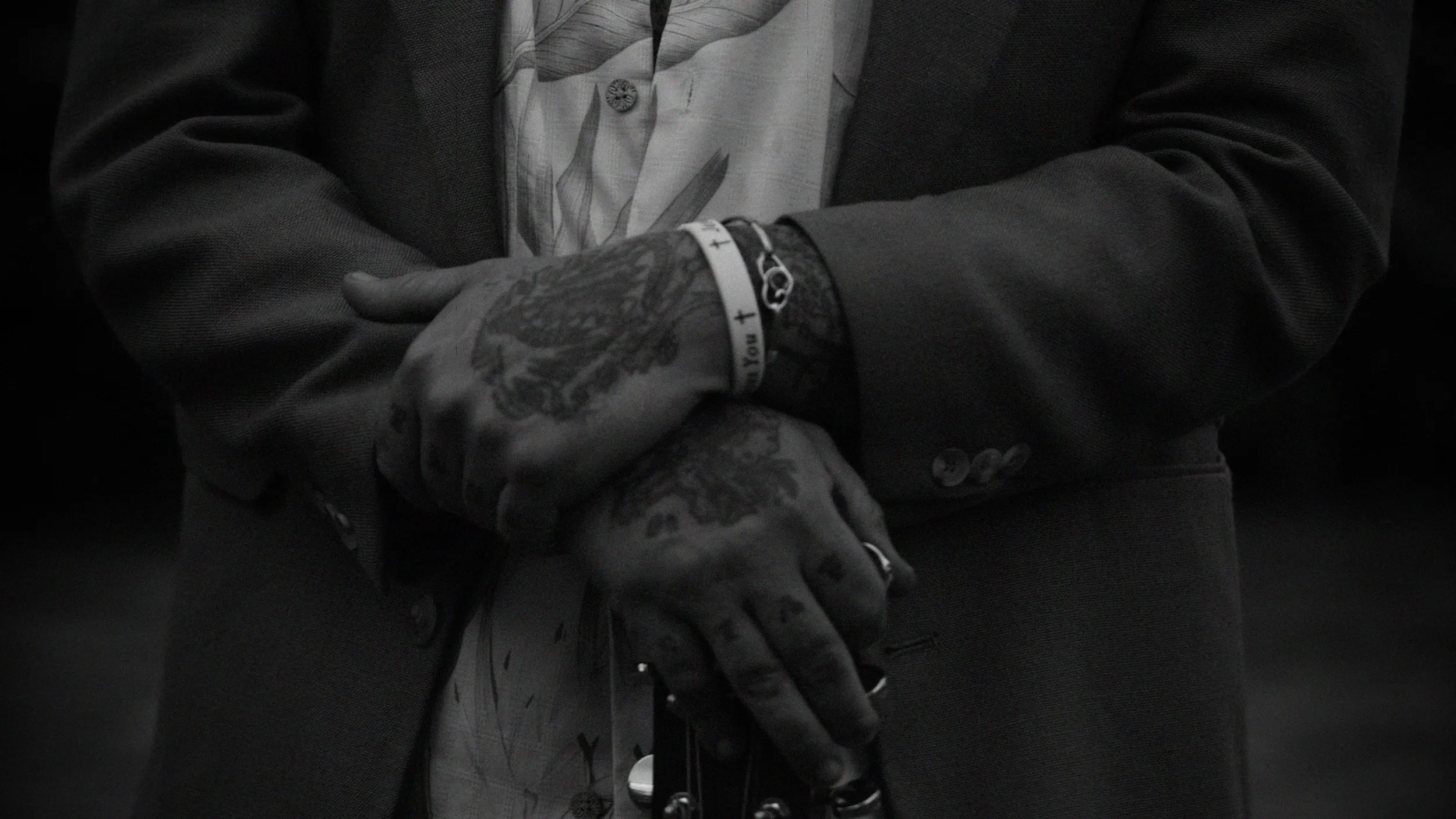 Black and white detail of tattooed hands clasped in front of a dark jacket.